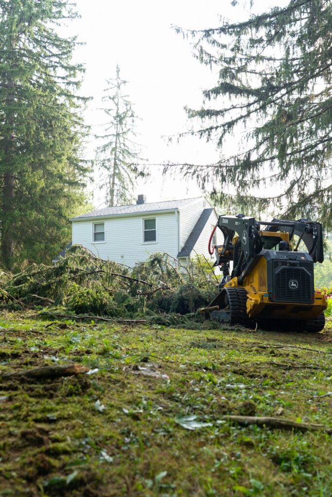 Tracked loader clearing a felled evergreen from a lawn beside a white house